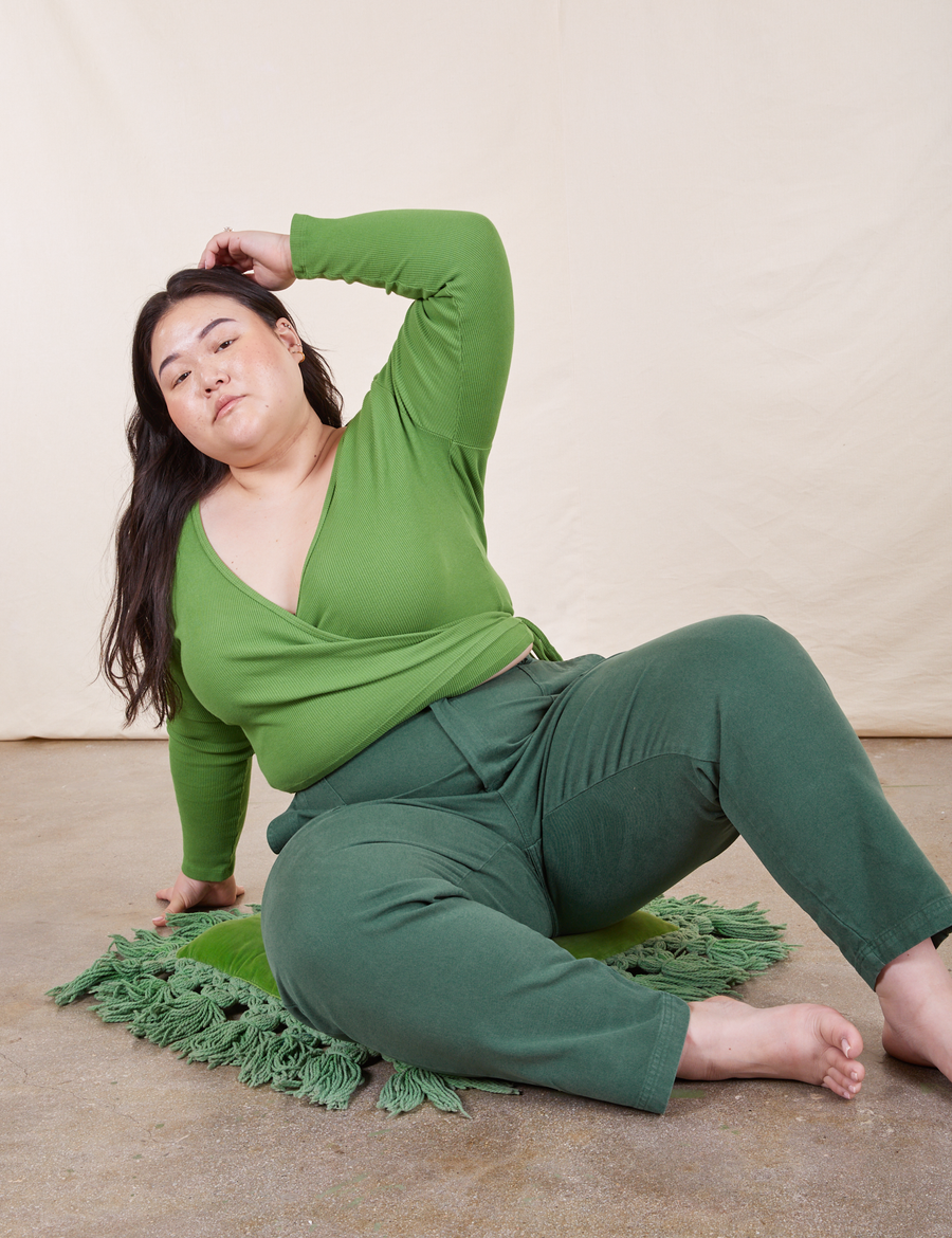 Ashley is sitting on a green cushion wearing Pencil Pants in Dark Emerald Green paired with a bright olive Wrap Top