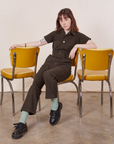 Hana is sitting on a yellow chair wearing Petite Short Sleeve Jumpsuit in Espresso Brown