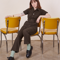 Hana is sitting on a yellow chair wearing Petite Short Sleeve Jumpsuit in Espresso Brown