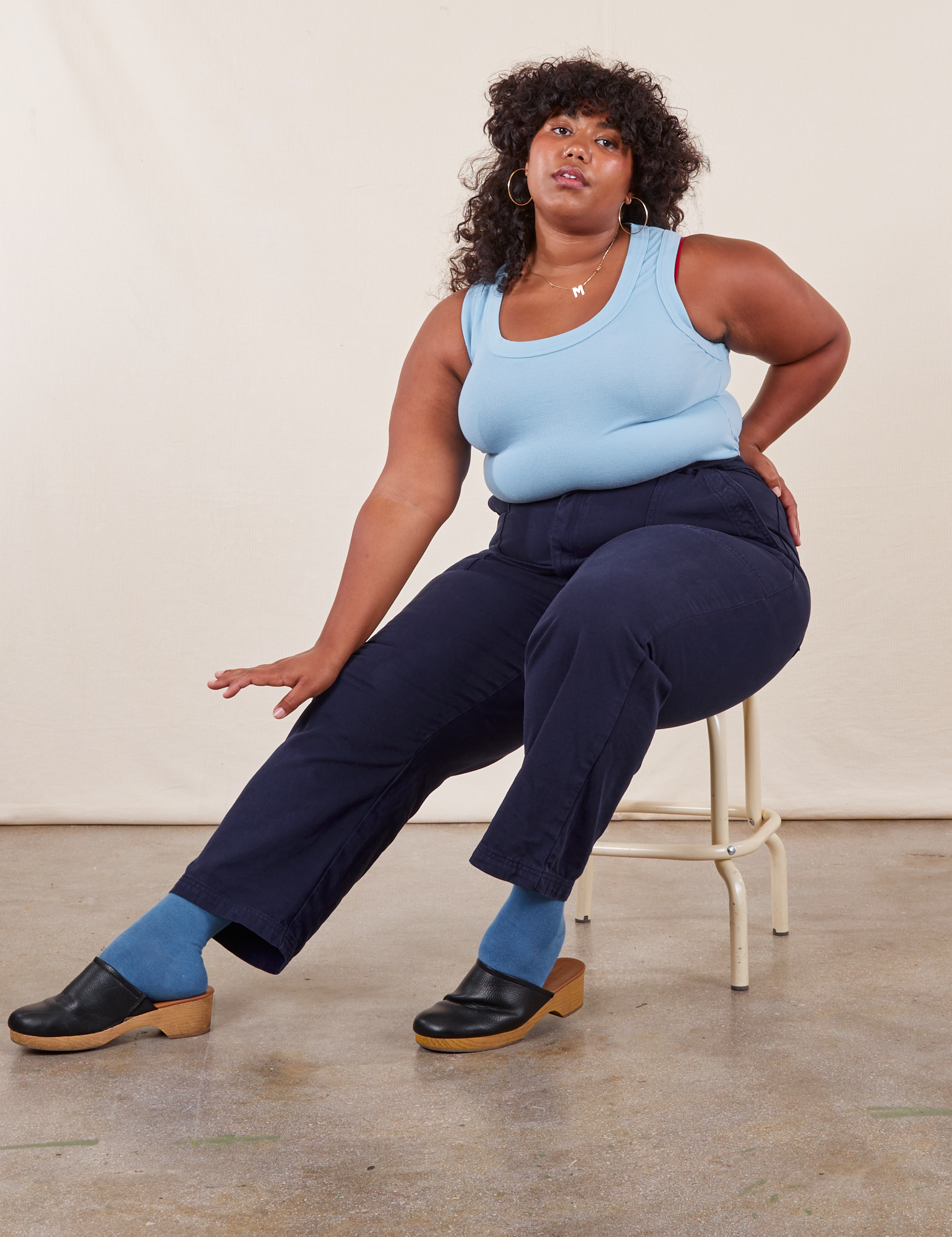 Morgan is sitting on a vintage stool wearing Work Pants in Navy Blue and a baby blue Tank Top