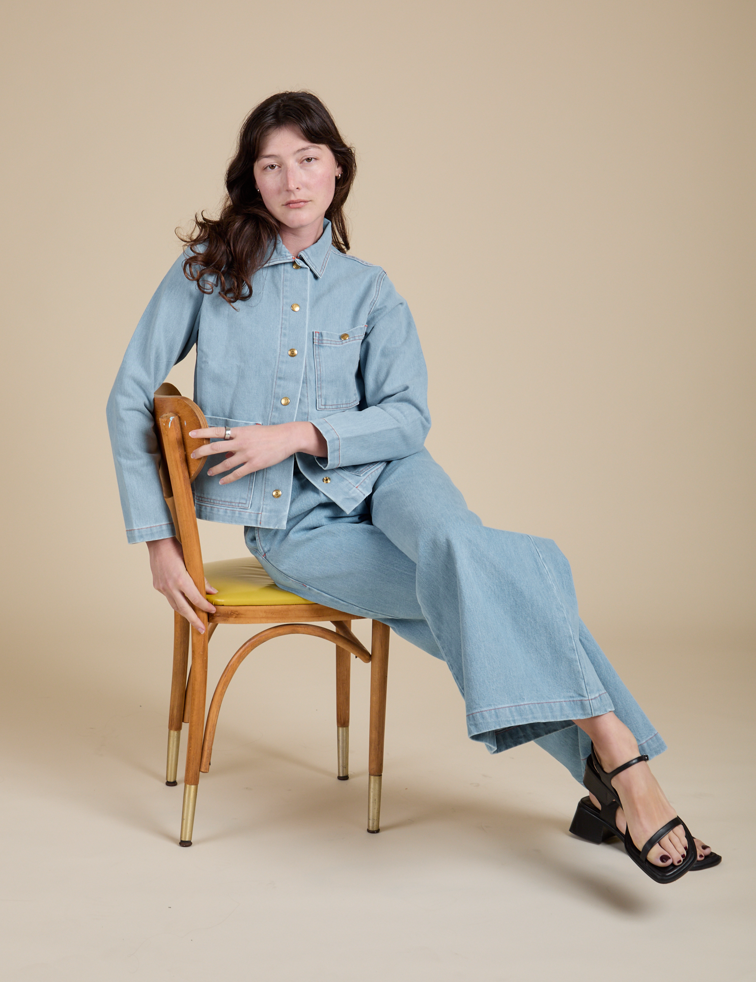 Alex is sitting on a chair and wearing Indigo Denim Work Jacket in Light Wash and light wash Indigo Wide Leg Trousers