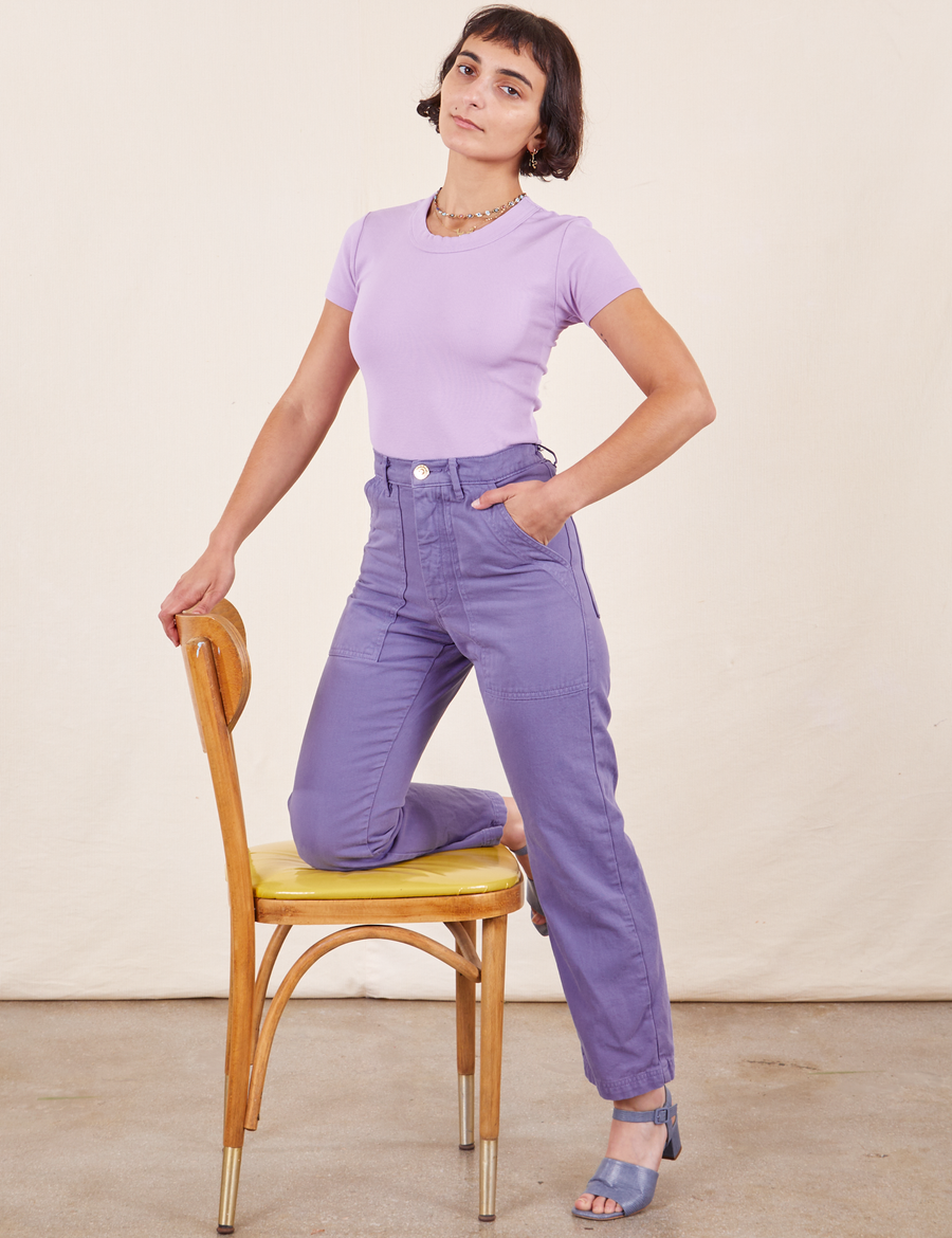 Work Pants in Faded Grape on Soraya wearing lilac purple Baby Tee. One knee is on a wooden chair with the other leg standing on the ground.