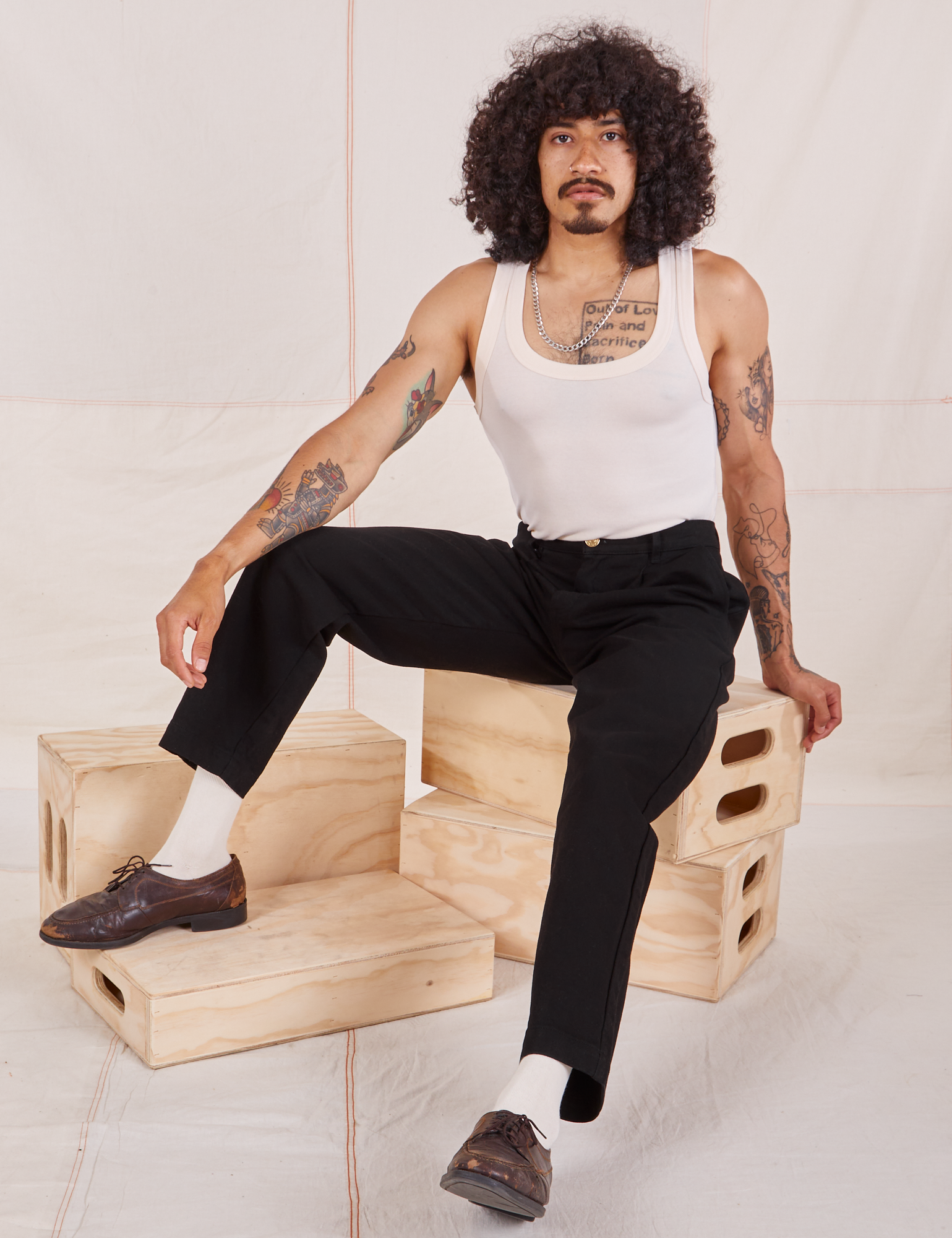 Jesse is sitting on a stack of wooden crates. They are wearing Denim Trouser Jeans in Black and a vintage off-white Tank Top