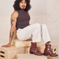 Jesse is sitting on a stack of wooden crates. They are wearing Heavyweight Trousers in Vintage Off-White and espresso brown Sleeveless Turtleneck.