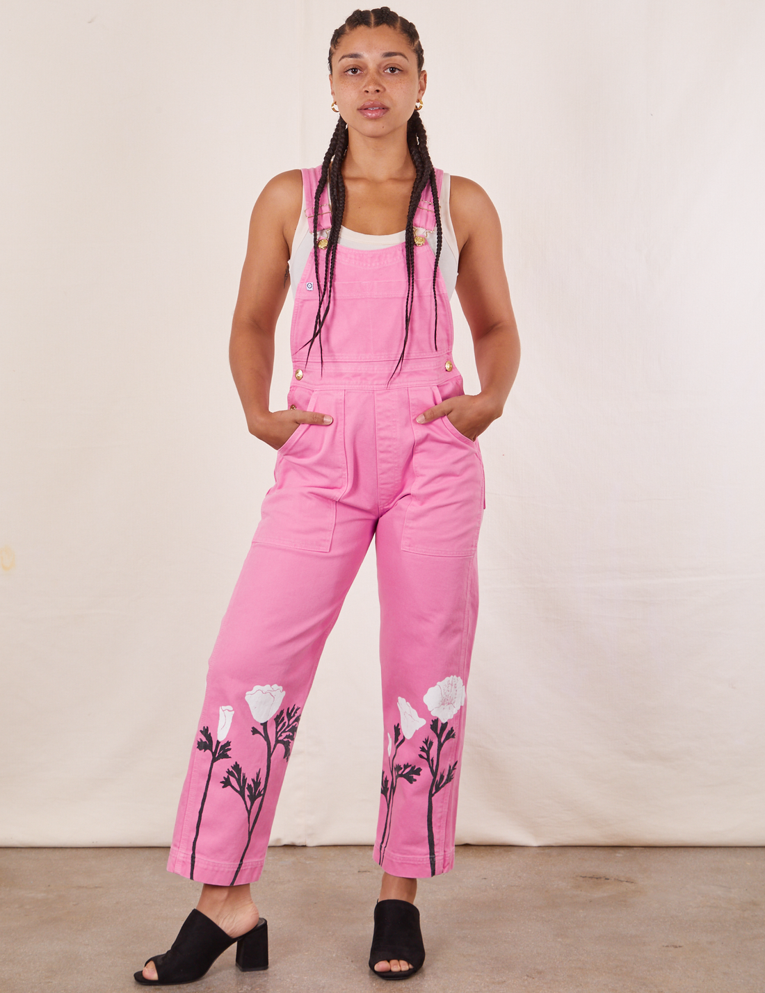 Gabi is 5'7" and wearing XXS California Poppy Overalls in Bubblegum Pink with a vintage off-white Tank Top underneath. She has both hands in the pockets.
