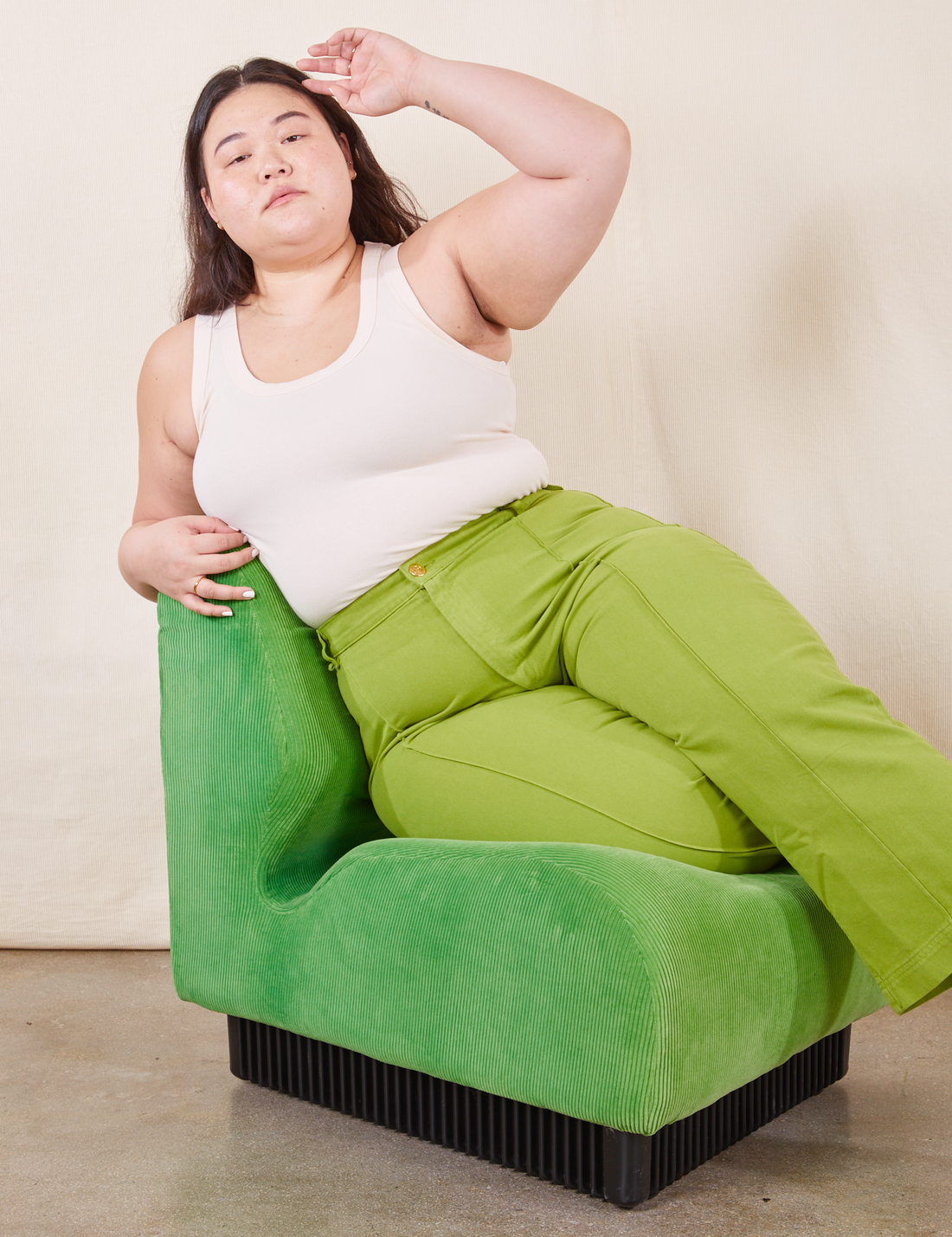 Ashley is sitting on a green upholstered chair wearing Western Pants in Gross Green paired with a vintage off-white Tank Top