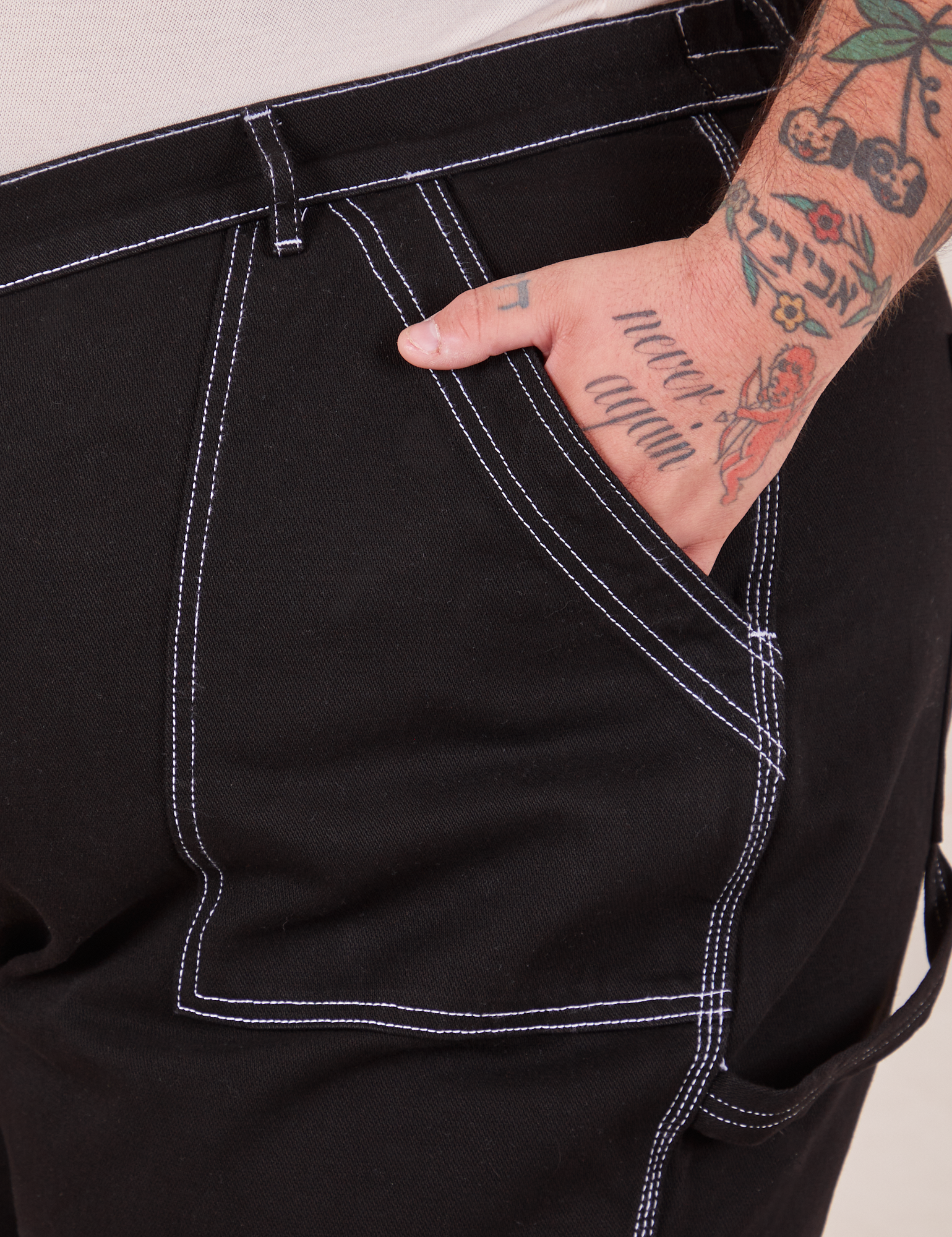 Front pocket close up of Carpenter Jeans in Black. Contrast white top stitching along edges of pocket. Sam has their hand in the pocket.