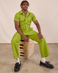 Issac is wearing Short Sleeve Jumpsuit in Gross Green and sitting on a stack of wooden crates