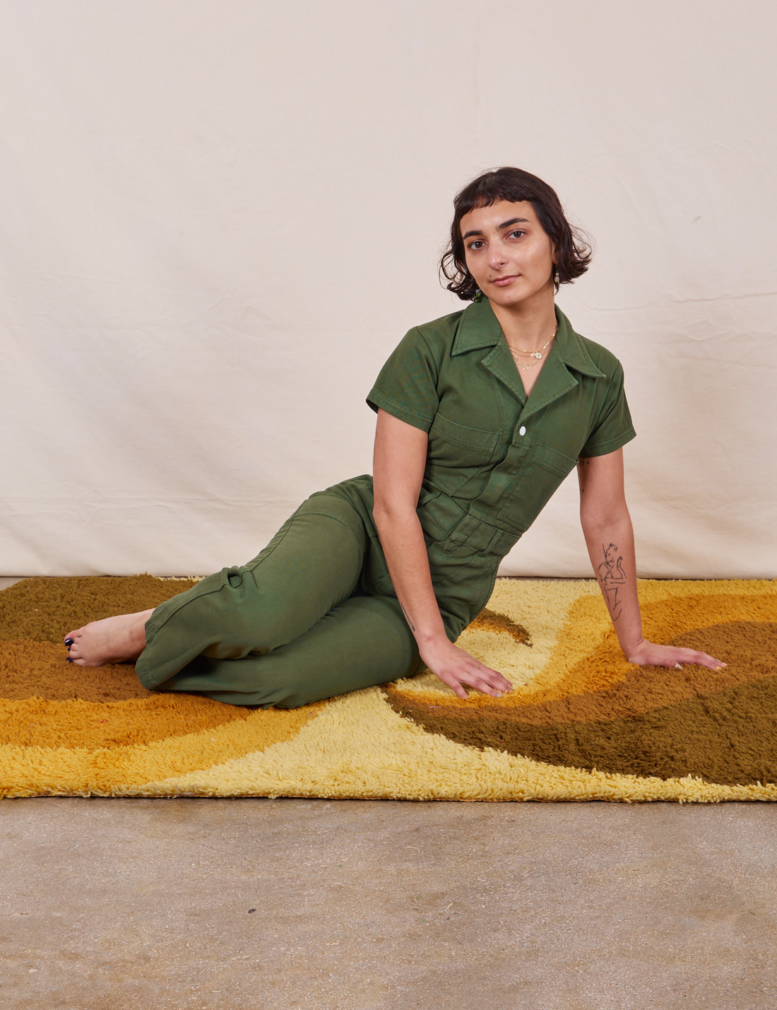 Soraya is sitting on a yellow rug wearing Petite Short Sleeve Jumpsuit in Dark Emerald Green