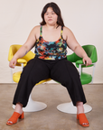 Ashley is sitting in between a yellow and green vintage chair. She is wearing Cropped Cami in Rainbow Magic Waters paired with black Petite Western Pants