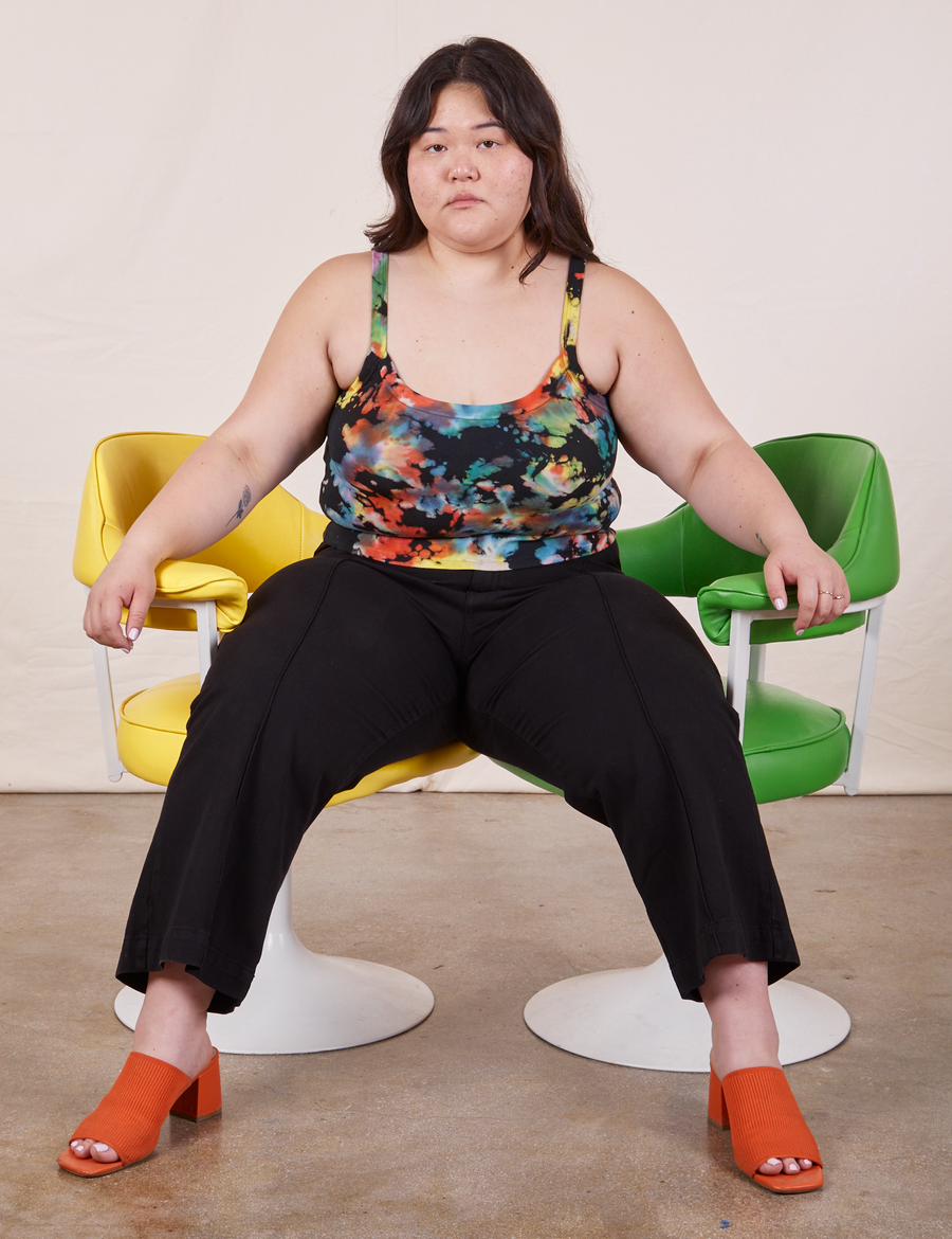 Ashley is sitting in between a yellow and green vintage chair. She is wearing Cropped Cami in Rainbow Magic Waters paired with black Petite Western Pants