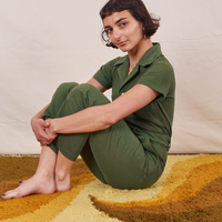Soraya is sitting on a yellow rug wearing Petite Short Sleeve Jumpsuit in Dark Emerald Green
