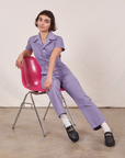 Soraya is sitting in a pink chair wearing Petite Short Sleeve Jumpsuit in Faded Grape