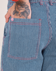 Back pocket close up of Railroad Stripe Denim Work Pants. Jesse has their hand in the pocket. Contrast red stitching and white and red sun baby logo tag on the edge of the pocket.