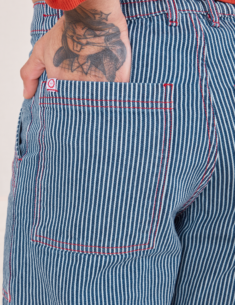 Back pocket close up of Railroad Stripe Denim Work Pants. Jesse has their hand in the pocket. Contrast red stitching and white and red sun baby logo tag on the edge of the pocket.