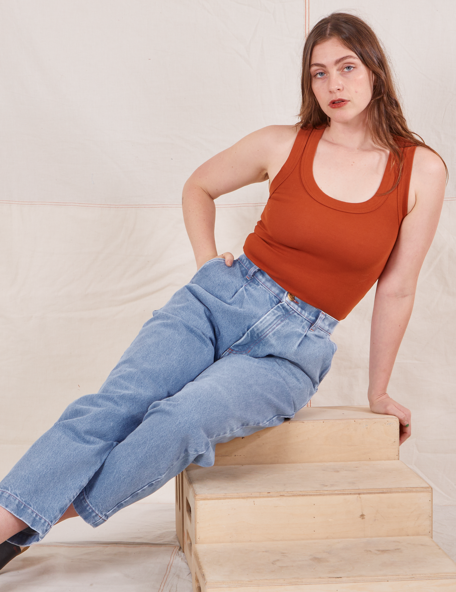 Allison is sitting on a wooden crate wearing Denim Trouser Jeans in Light Wash and a burnt orange Tank Top