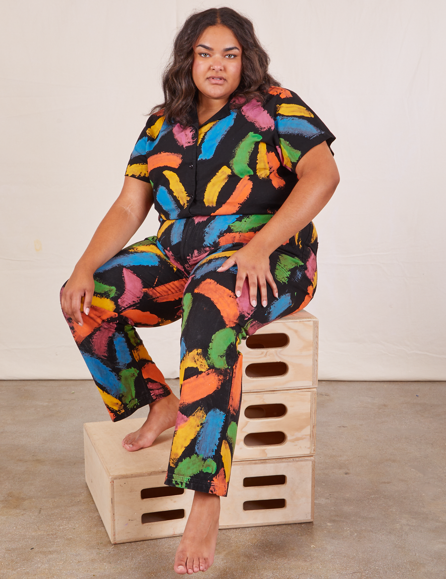 Alicia is sitting on a stack of wooden crates wearing Pantry Button Up in Paint Stroke and matching Work Pants