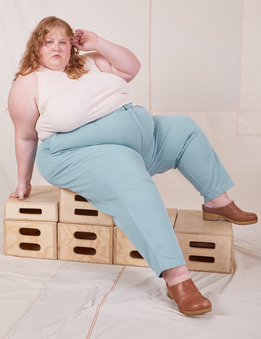 Catie is sitting on a stack of wooden crates. She is wearing Heavyweight Trousers in Baby Blue and vintage off-white Sleeveless Turtleneck