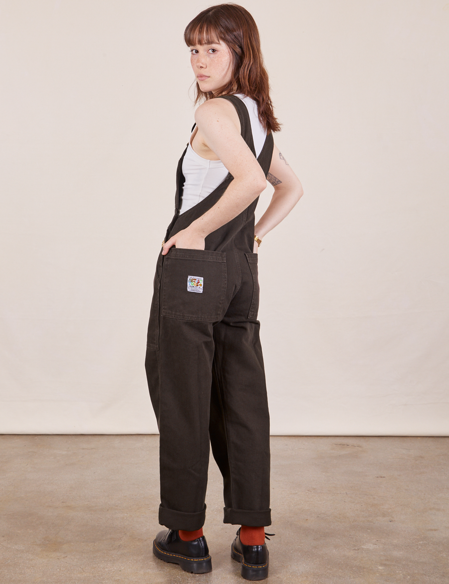 Angled back view of Original Overalls in Mono Espresso. Hana has both hands in the back pockets.