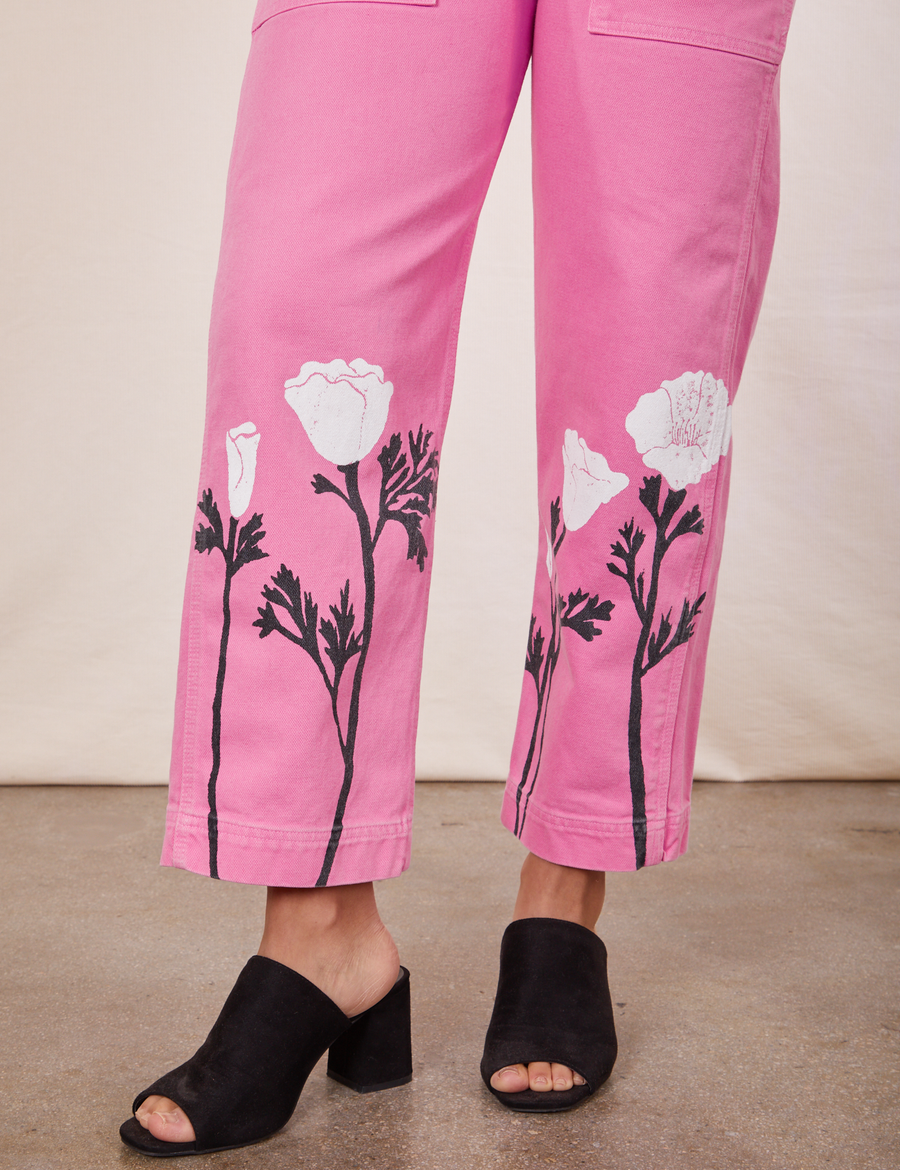 Pant leg close up of California Poppy Overalls in Bubblegum Pink worn by Gabi. Paintstamped white poppies with black stems and leaves along the bottom.