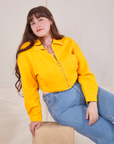 Sydney is sitting on a wooden crate wearing the Ricky Jacket in Sunshine Yellow