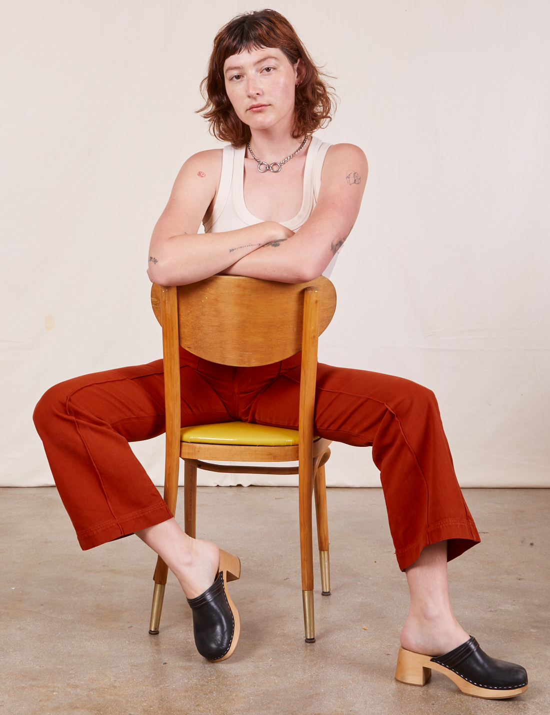 Alex is sitting backwards in a wooden chair wearing Western Pants in Paprika and a vintage off-white Tank Top