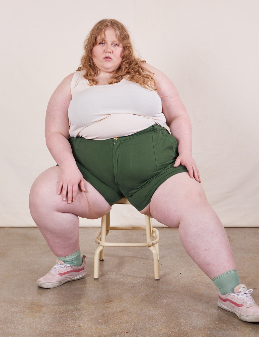 Catie is sitting on a vintage stool wearing Classic Work Shorts in Dark Emerald Green paired with a vintage off-white Tank Top