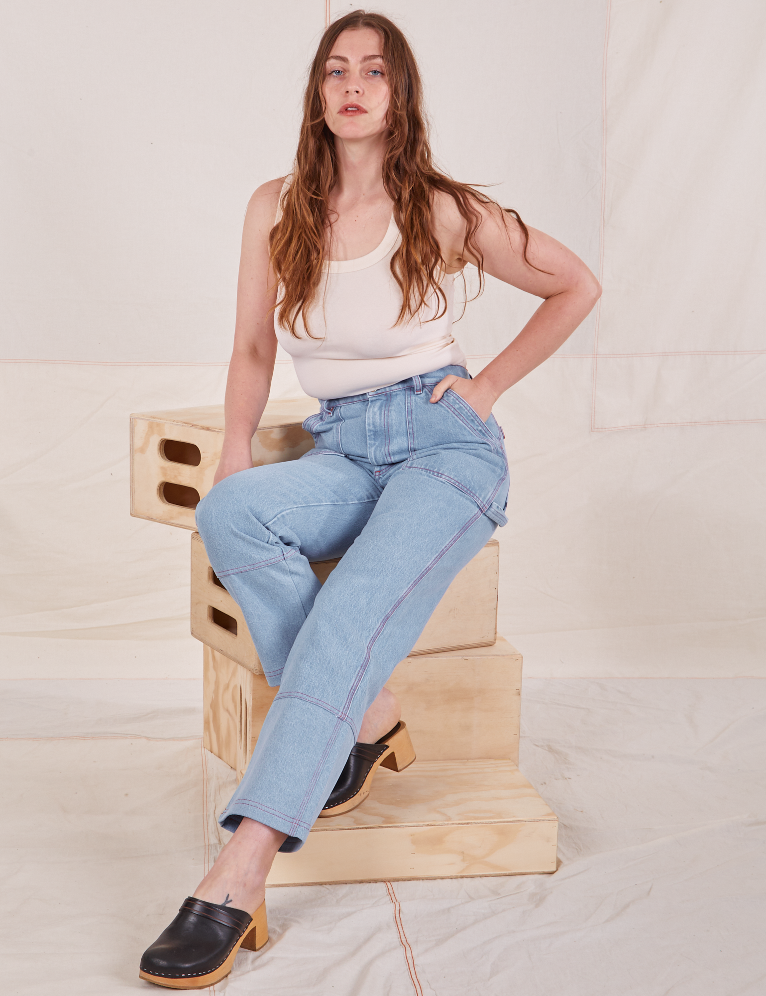 Allison is sitting on a stack of wooden crates. She is wearing Carpenter Jeans in Light Wash paired with a vintage off-white Tank Top.