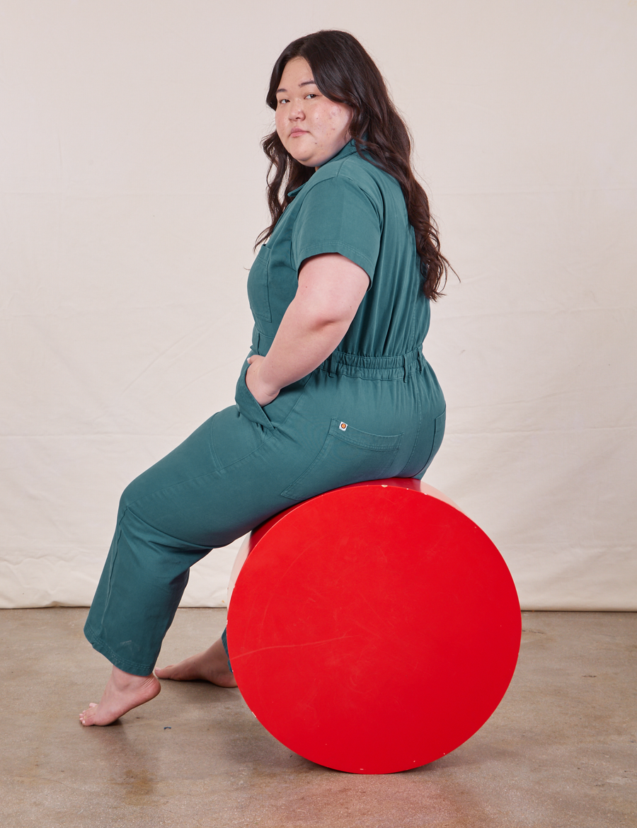 Ashley is sitting on a red circular platform wearing Petite Short Sleeve Jumpsuit in Marine Blue