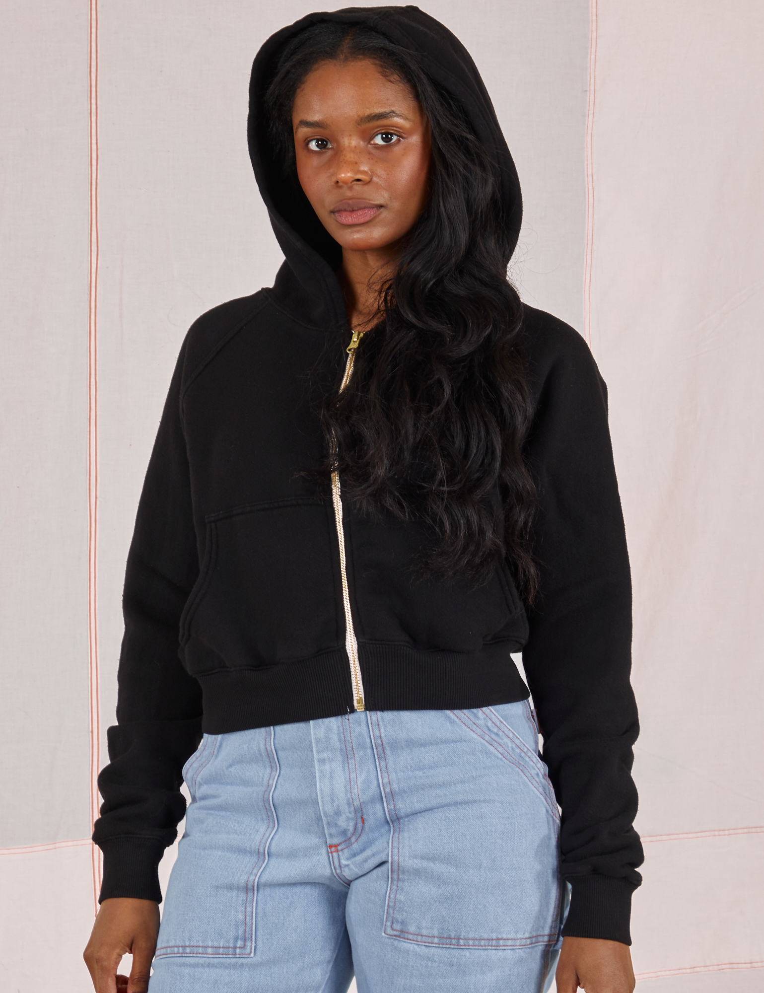 Kandia is wearing Cropped Zip Hoodie in Basic Black with the hood up and light wash Carpenter Jeans