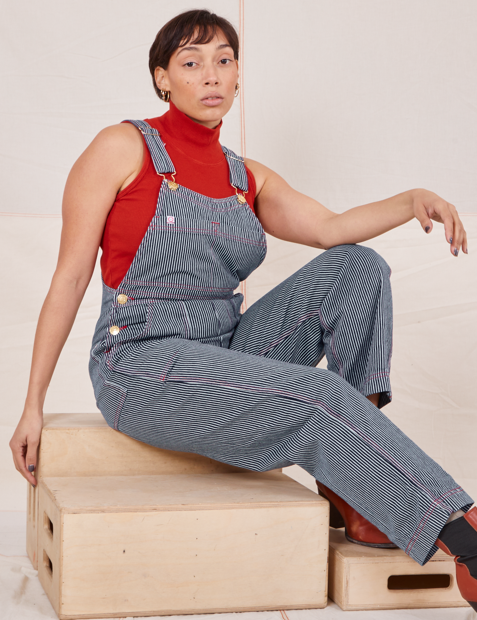 Taira is sitting on a wooden crate wearing Railroad Stripe Denim Original Overalls and a paprika Sleeveless Turtleneck