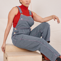 Taira is sitting on a wooden crate wearing Railroad Stripe Denim Original Overalls and a paprika Sleeveless Turtleneck