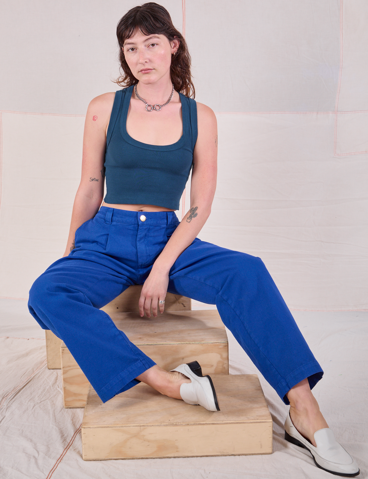 Alex is sitting on a wooden crate wearing Mid-Rise Pleated Trousers in Royal Blue and lagoon Cropped Tank