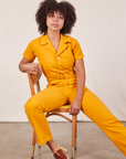 Gabi is sitting in a chair wearing Short Sleeve Jumpsuit in Mustard Yellow