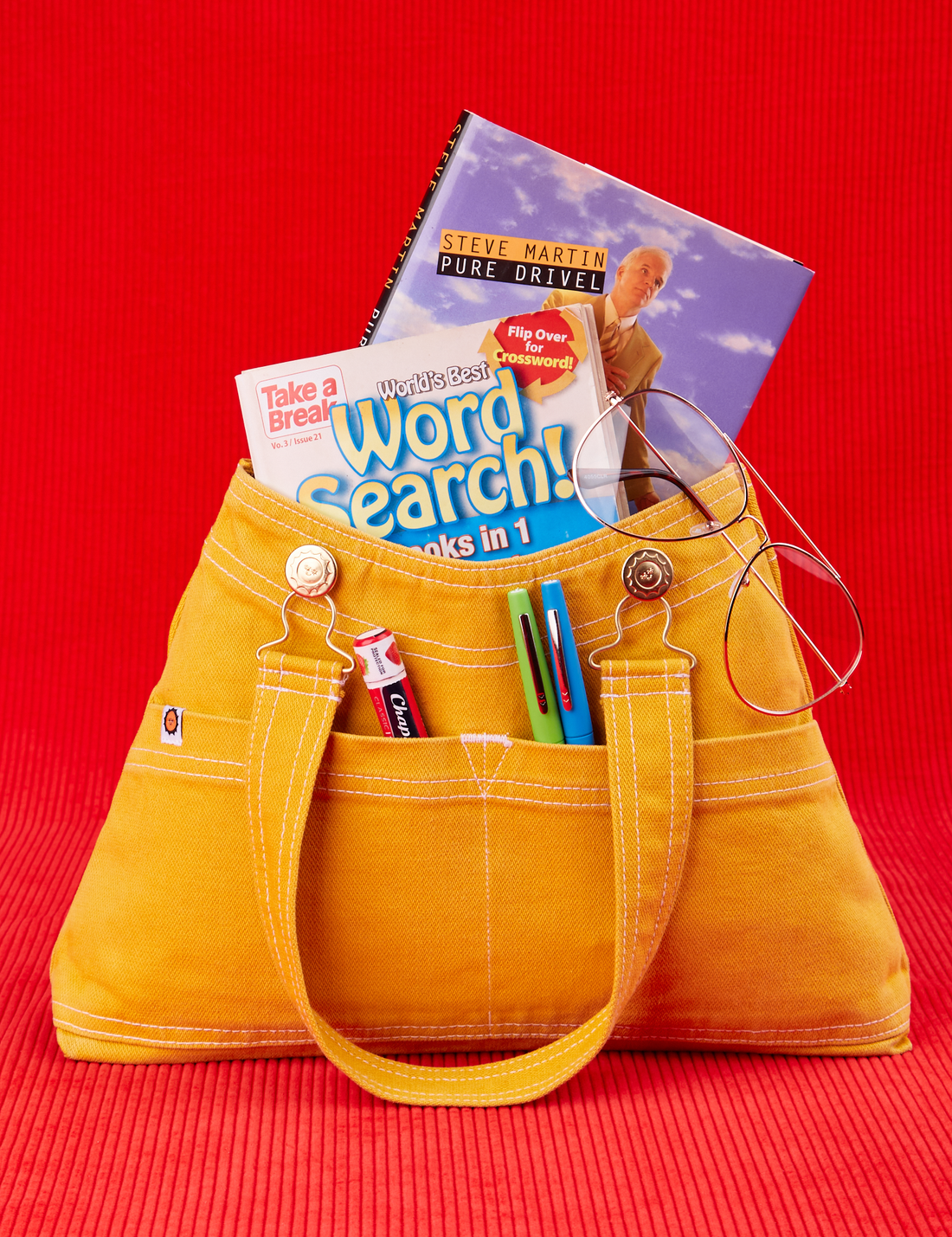 Overall Handbag in Mustard Yellow with two books inside main compartment of bag. Chapstick and two pens are in the outer pockets. Glasses hung along edge of bag.
