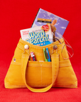 Overall Handbag in Mustard Yellow with two books inside main compartment of bag. Chapstick and two pens are in the outer pockets. Glasses hung along edge of bag.