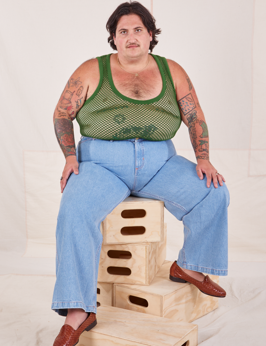 Sam is sitting on a stack of wooden crates. They are wearing Mesh Tank Top in Lawn Green and light wash Sailor Jeans
