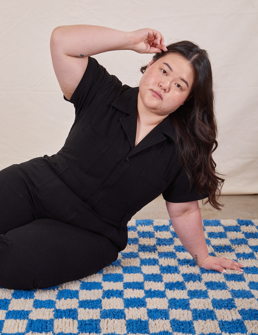 Ashley is sitting on a blue and white checkered rug wearing Petite Short Sleeve Jumpsuit in Basic Black