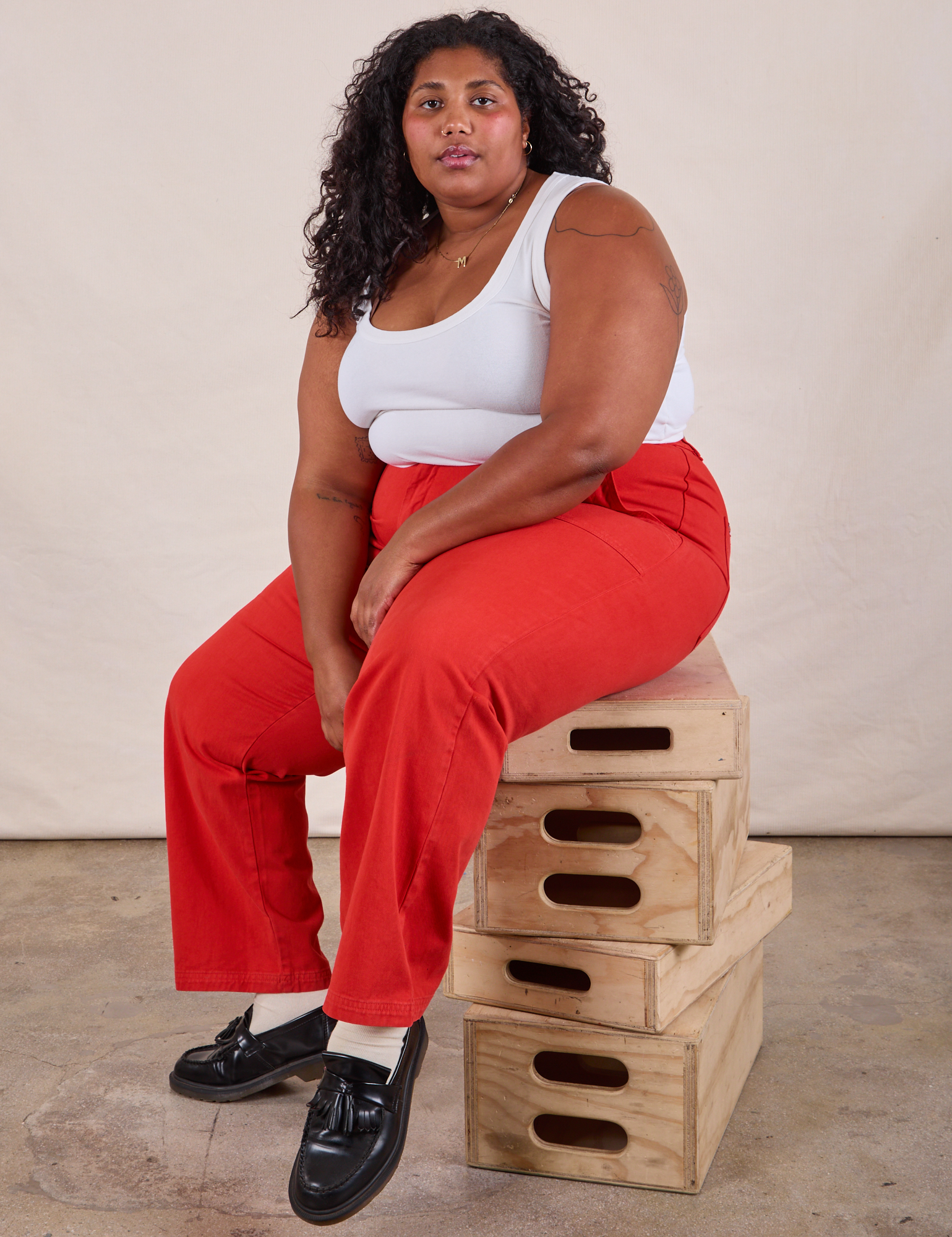 Morgan is sitting on a stack of wooden crates wearing Work Pants in Mustang Red