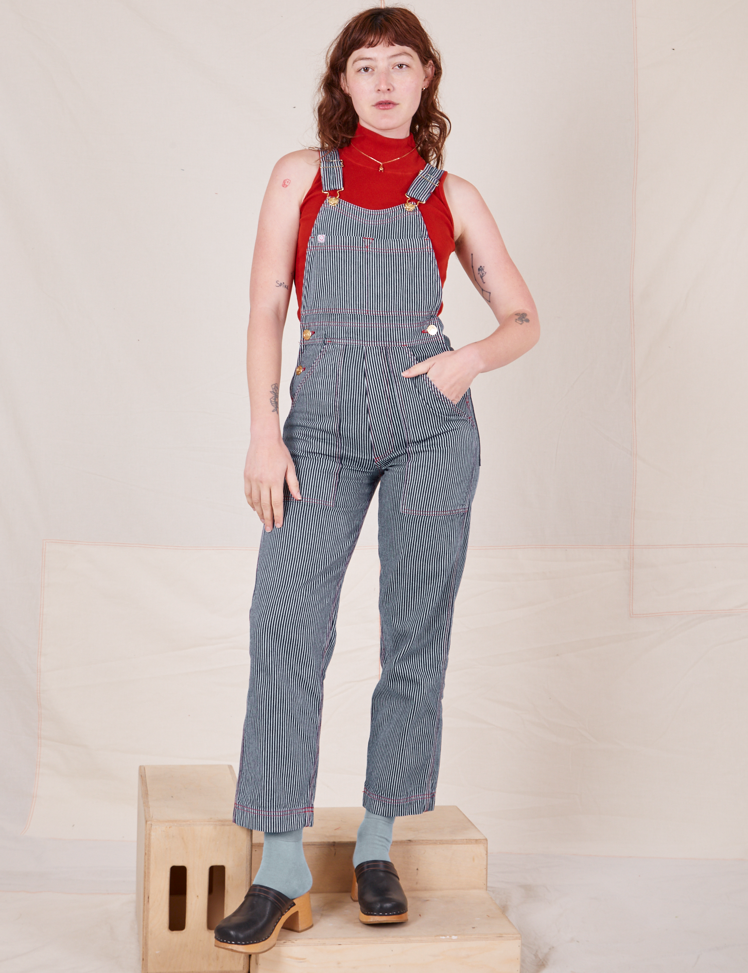 Alex is 5&#39;8&quot; and wearing P Railroad Stripe Denim Original Overalls paired with paprika Sleeveless Turtleneck