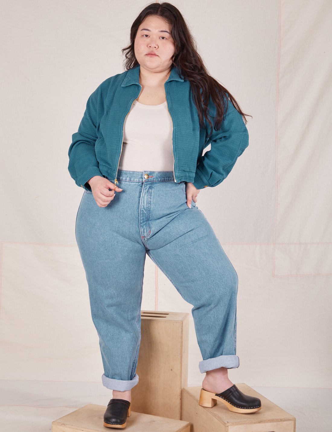 Ashley is standing on wooden crates wearing the Ricky Jacket in Marine Blue, a vintage off-white Tank Top underneath and light wash Frontier Jeans