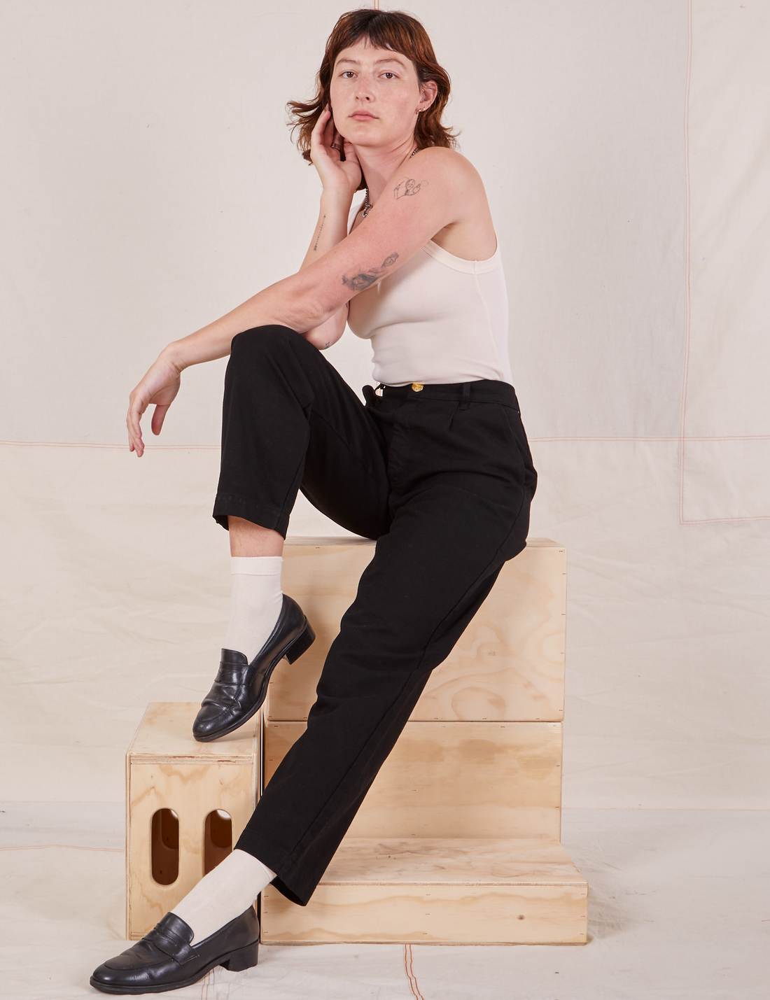 Alex is sitting on a stack of wooden crates wearing Denim Trouser Jeans in Black and a vintage off-white Tank Top