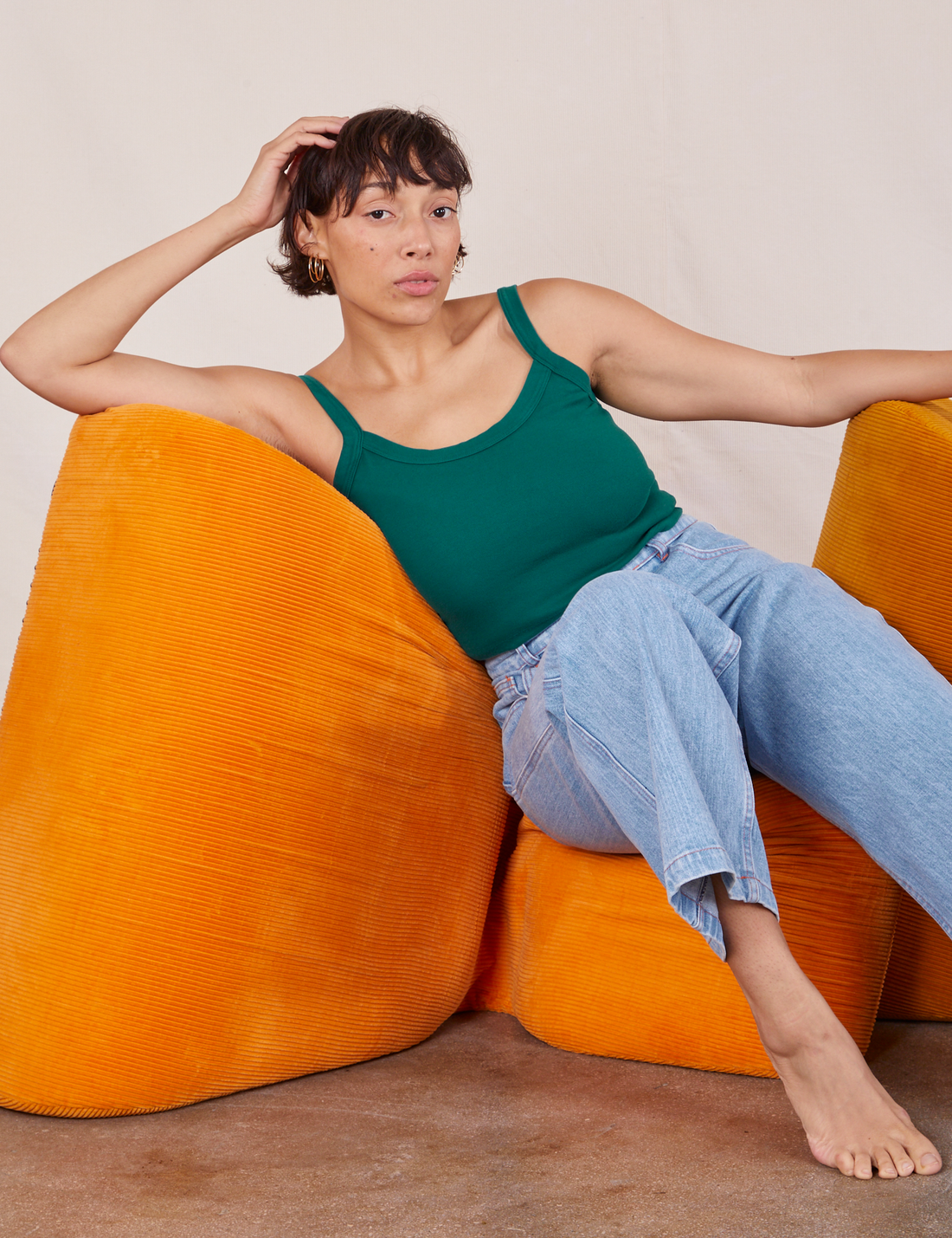 Tiara is sitting on an orange upholstered chair wearing Cropped Cami in Hunter Green and light wash Frontier Jeans