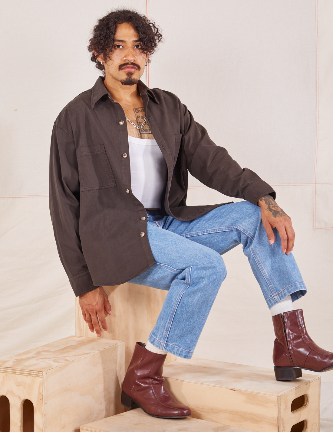 Jesse is sitting on a wooden crate. They are wearing Oversize Overshirt in Espresso Brown, vintage off-white Cropped Tank Top and light wash Frontier Jeans