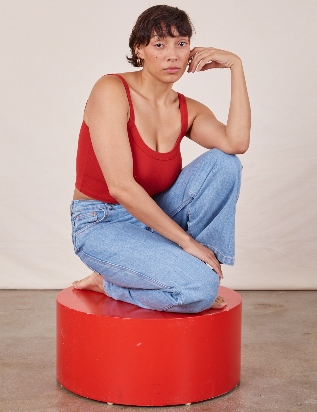 Tiara is kneeling on a red circular platform wearing Cropped Cami in Mustang Red and light wash Western Pants