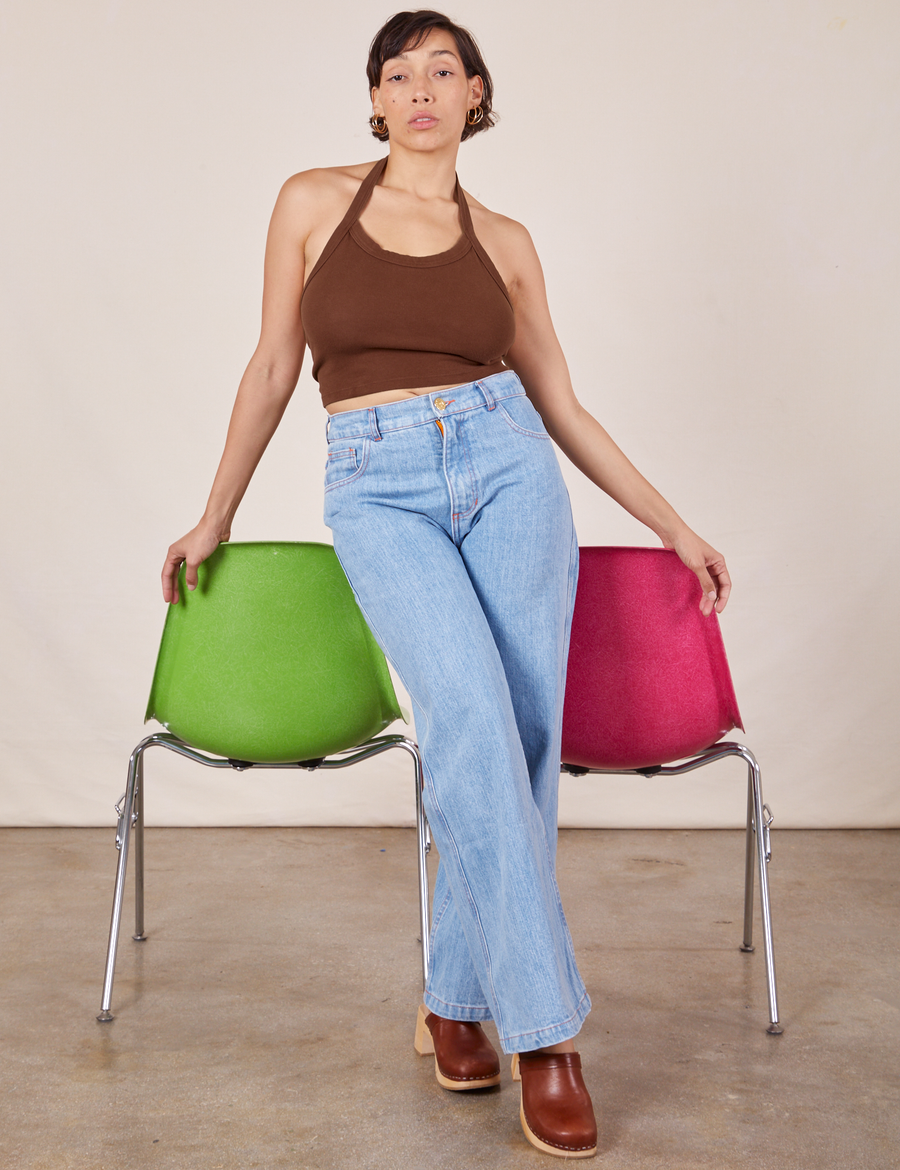 Tiara is standing in front of a green and pink chair with her hands resting on each. She is wearing Halter Top in Fudgesicle Brown and light wash Sailor Jeans.