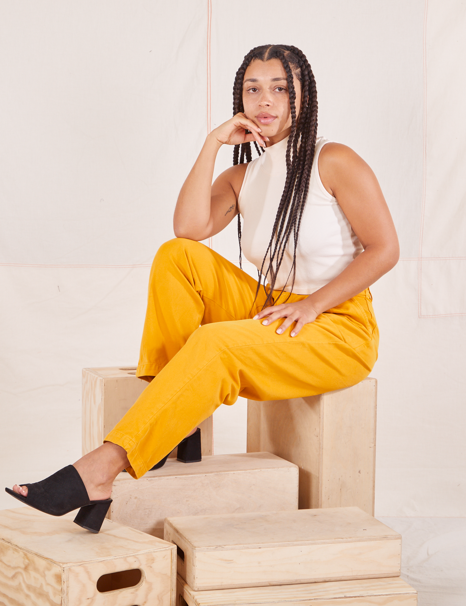 Gabi is sitting on a wooden crate wearing Organic Trousers in Mustard Yellow