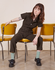 Hana is sitting in a yellow chair wearing Petite Short Sleeve Jumpsuit in Espresso Brown