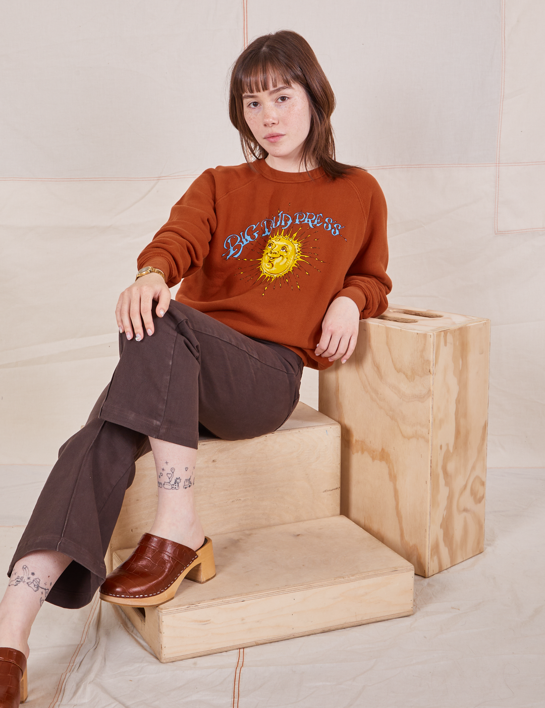 Hana is sitting on a wooden crate wearing Bill Ogden's Sun Baby Crew and espresso brown Western Pants