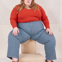 Catie is sitting on a wooden crate wearing Railroad Stripe Denim Work Pants and paprika Long Sleeve V-Neck Tee